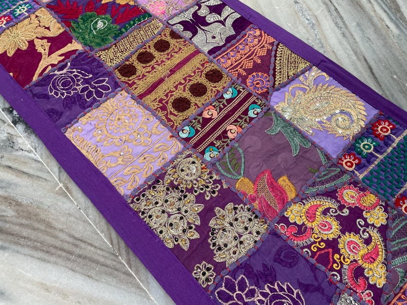 May include: A patchwork table runner with a deep purple border. The runner is composed of various fabric squares in shades of purple, gold, and pink, featuring embroidered floral and paisley designs. The runner is approximately 60 inches long.