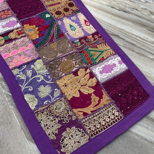 May include: A rectangular, patchwork table runner with a purple border and a tassel. The patchwork design features squares of various colors and patterns, including gold embroidery, floral designs, and paisley motifs.
