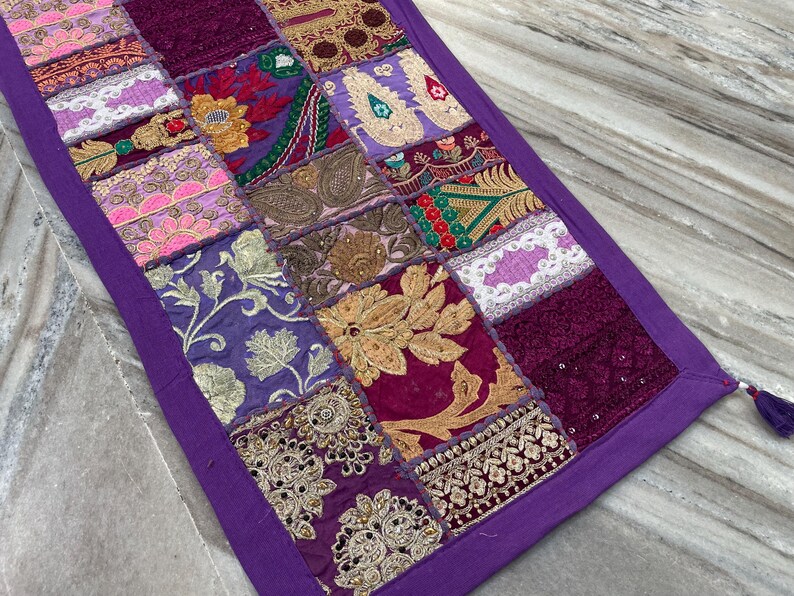 May include: A rectangular, patchwork table runner with a purple border and a tassel. The patchwork design features squares of various colors and patterns, including gold embroidery, floral designs, and paisley motifs.