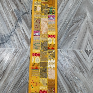May include: A long, rectangular yellow patchwork textile with a variety of patterned squares. The squares feature floral, geometric, and abstract designs in shades of yellow, gold, green, and red. The edges are bordered with a solid yellow fabric.
