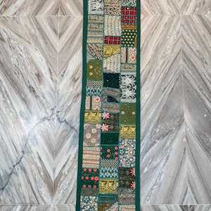 May include: A long, rectangular, patchwork textile in shades of green, beige, and red. The design features a variety of patterned squares and a dark green border with tassels at each end. This decorative piece is likely a table runner or wall hanging.
