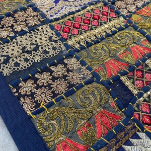 May include: Close-up of a patchwork textile with intricate embroidery. The design features squares of navy blue, gold, red, and cream-colored fabrics. The embroidery includes floral and geometric patterns, with gold sequins and thread.