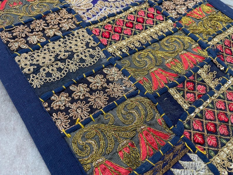 May include: Close-up of a patchwork textile with intricate embroidery. The design features squares of navy blue, gold, red, and cream-colored fabrics. The embroidery includes floral and geometric patterns, with gold sequins and thread.