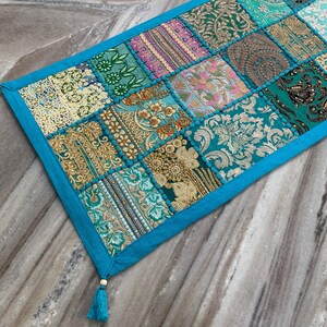 May include: A rectangular patchwork table runner with a teal border and tassels. The runner features a variety of patterned fabric squares in teal, gold, pink, and green. The design includes floral and geometric patterns.