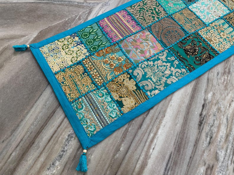 May include: A rectangular patchwork table runner with a teal border and tassels. The runner features a variety of patterned fabric squares in teal, gold, pink, and green. The design includes floral and geometric patterns.