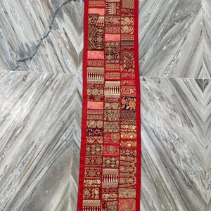 Op de afbeelding: Een rechthoekig, rood textiel met een patchwork-ontwerp. Het patchwork bevat verschillende patronen in tinten rood, goud en roze. Het textiel is waarschijnlijk een decoratief item, zoals een tafelloper of wandkleed.