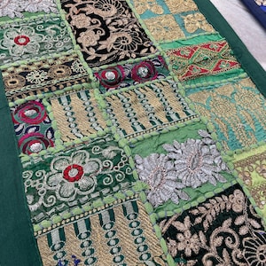 May include: A patchwork textile with a green border. The design features squares of various embroidered fabrics in shades of green, gold, black, and red. Floral and geometric patterns are prominent, creating a textured and colorful display.