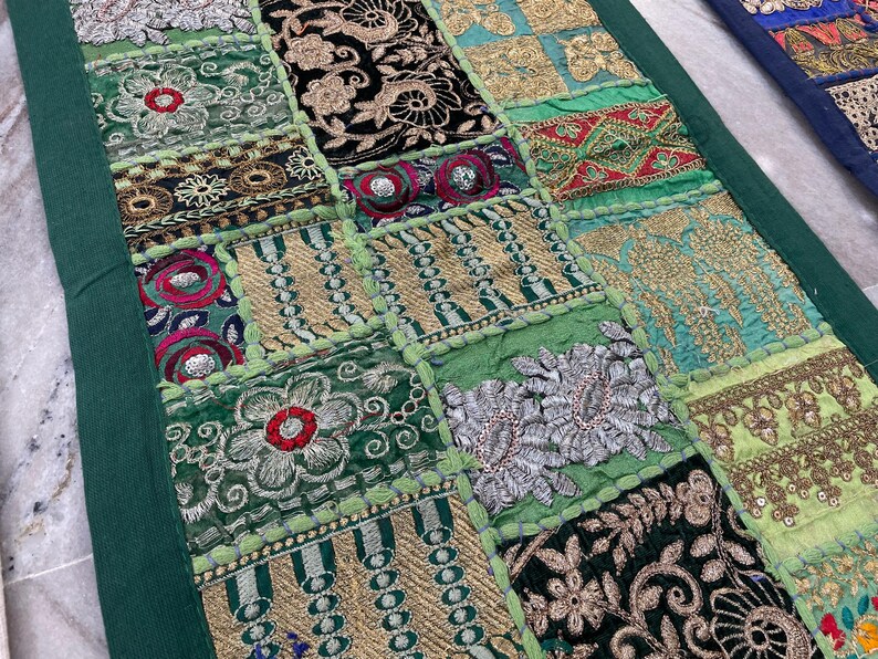 May include: A patchwork textile with a green border. The design features squares of various embroidered fabrics in shades of green, gold, black, and red. Floral and geometric patterns are prominent, creating a textured and colorful display.