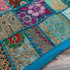 May include: Close-up of a patchwork textile with a vibrant mix of colors and patterns. The design features squares of embroidered fabric in teal, gold, red, and purple. The edges are outlined with a bright turquoise border, creating a decorative and textured surface.