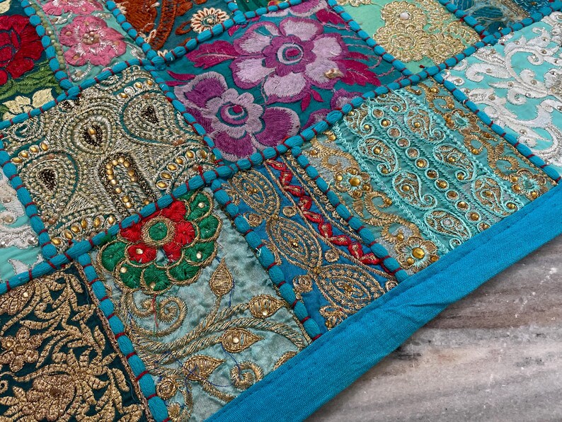 May include: Close-up of a patchwork textile with a vibrant mix of colors and patterns. The design features squares of embroidered fabric in teal, gold, red, and purple. The edges are outlined with a bright turquoise border, creating a decorative and textured surface.