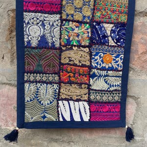 May include: A rectangular textile with a patchwork design. The patches feature various embroidered patterns in shades of blue, pink, gold, and green. The edges are trimmed with a dark blue border and tassels.