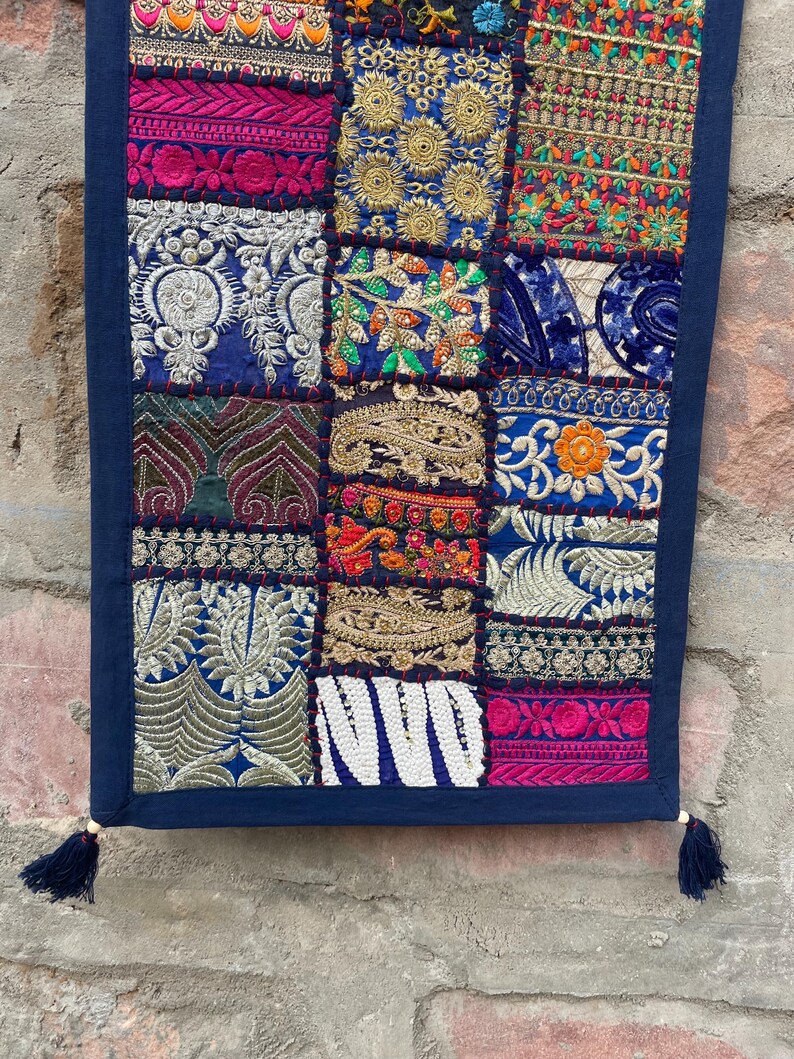 May include: A rectangular textile with a patchwork design. The patches feature various embroidered patterns in shades of blue, pink, gold, and green. The edges are trimmed with a dark blue border and tassels.