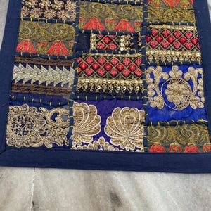 May include: A patchwork textile with a dark blue border. The design features a variety of embroidered squares in shades of gold, red, and blue. The squares have intricate patterns and textures, creating a rich, decorative piece.