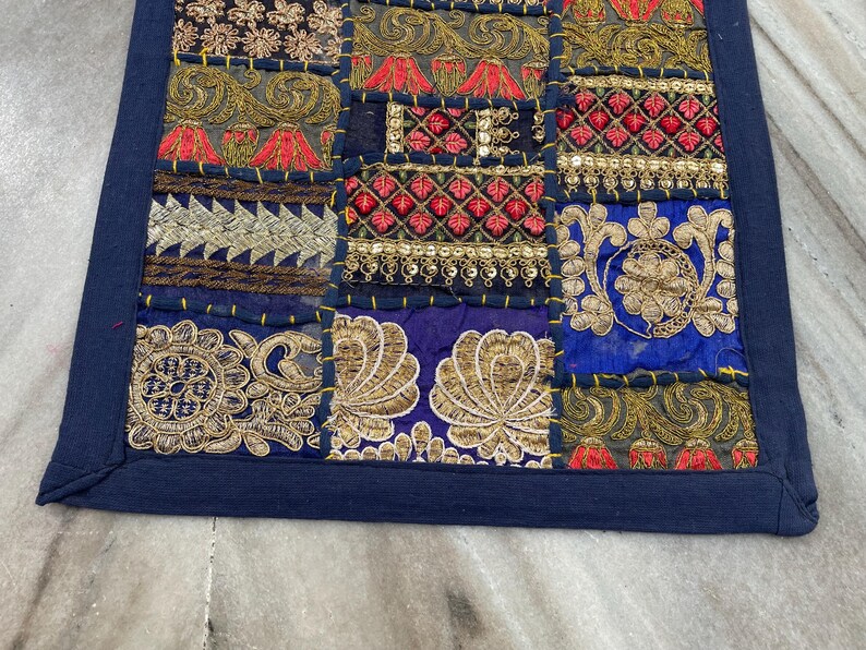 May include: A patchwork textile with a dark blue border. The design features a variety of embroidered squares in shades of gold, red, and blue. The squares have intricate patterns and textures, creating a rich, decorative piece.