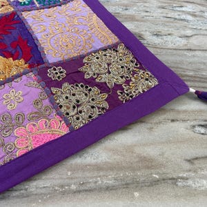 May include: A patchwork textile with a purple border and a tassel. The textile features embroidered squares in shades of purple, pink, gold, and red. The design includes floral and geometric patterns, with metallic thread accents.