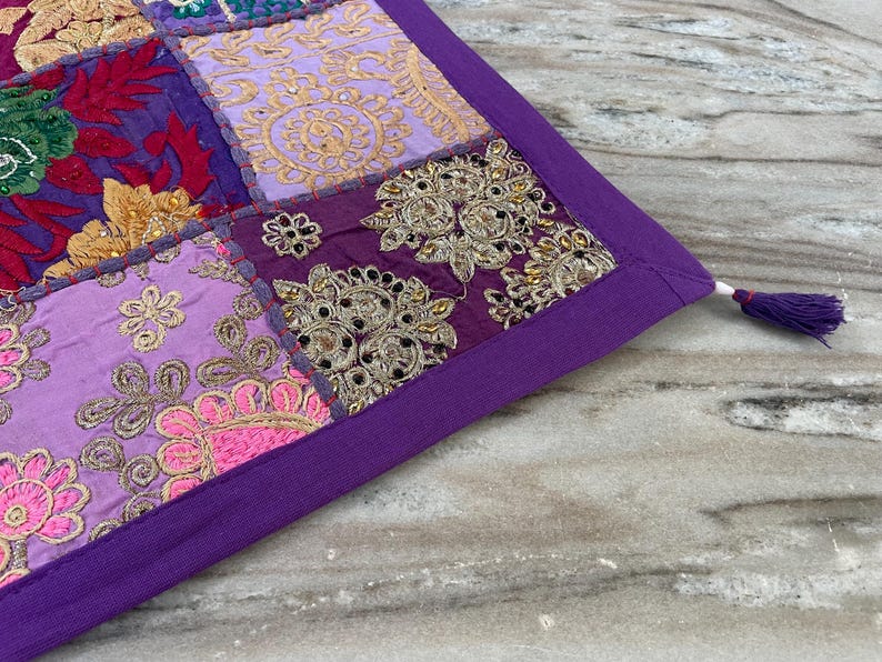 May include: A patchwork textile with a purple border and a tassel. The textile features embroidered squares in shades of purple, pink, gold, and red. The design includes floral and geometric patterns, with metallic thread accents.