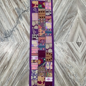 May include: A rectangular, patchwork table runner with a deep purple border and tassels. The runner is composed of various fabric squares in shades of purple, pink, gold, and green, featuring floral and paisley patterns.