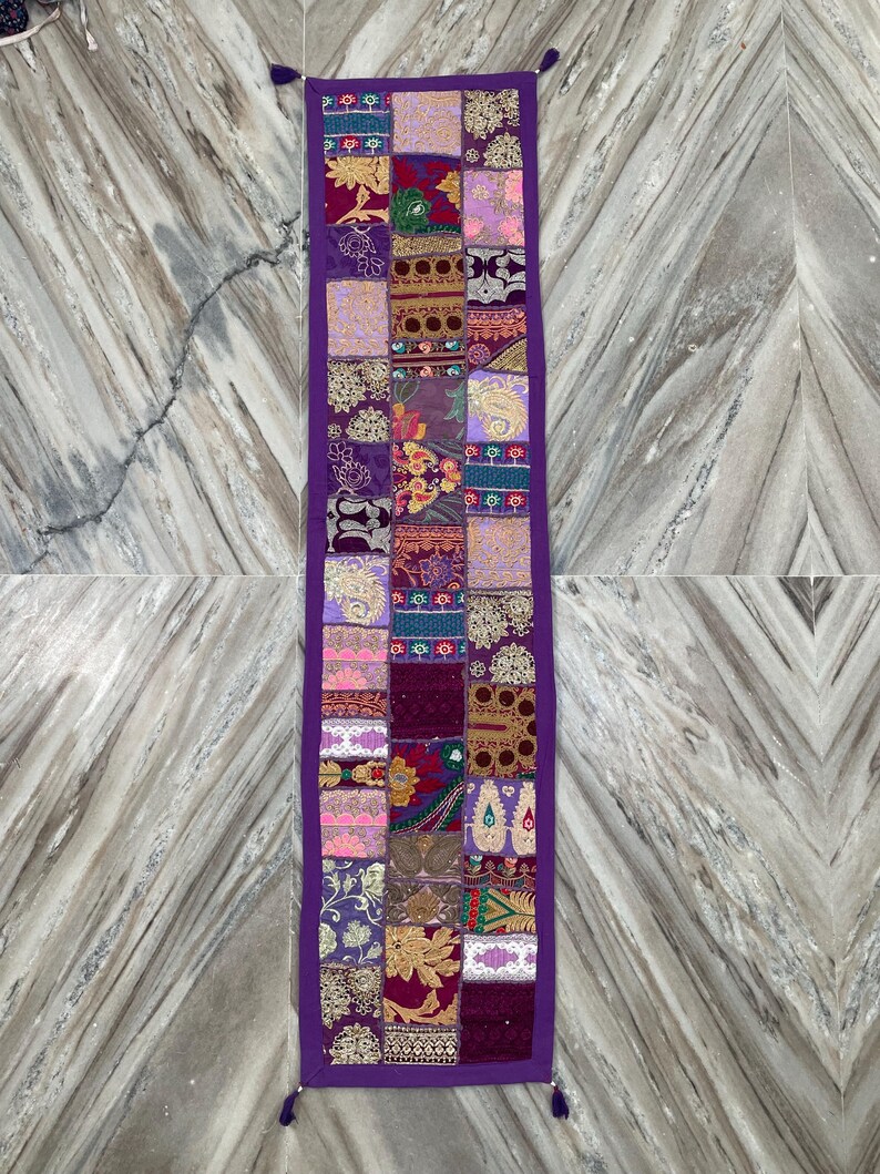 May include: A rectangular, patchwork table runner with a deep purple border and tassels. The runner is composed of various fabric squares in shades of purple, pink, gold, and green, featuring floral and paisley patterns.