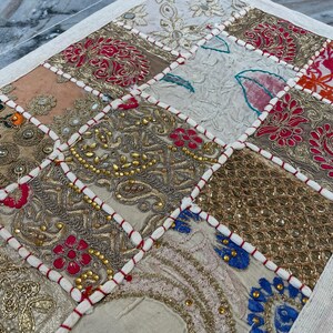 May include: A patchwork textile with a variety of embroidered squares in shades of gold, pink, red, and cream. The squares are outlined with white stitching and red thread, creating a decorative pattern.