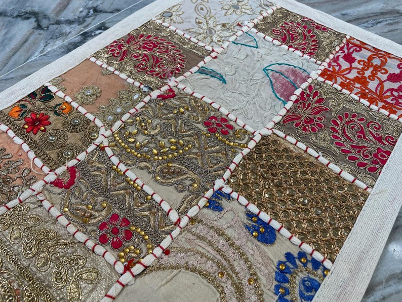 May include: A patchwork textile with a variety of embroidered squares in shades of gold, pink, red, and cream. The squares are outlined with white stitching and red thread, creating a decorative pattern.