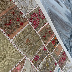 May include: A patchwork textile with a variety of colors and textures. The design features squares of fabric in gold, red, pink, and beige, with intricate embroidery and beadwork. The edges are finished with a white border.