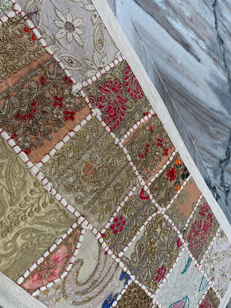 May include: A patchwork textile with a variety of colors and textures. The design features squares of fabric in gold, red, pink, and beige, with intricate embroidery and beadwork. The edges are finished with a white border.