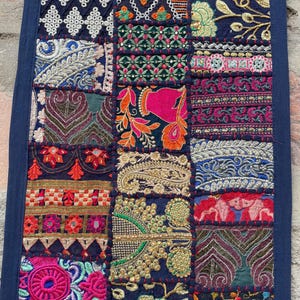 May include: A navy blue patchwork textile with intricate embroidery in various colors. The design features geometric patterns, floral motifs, and ornate details. The textile is rectangular and appears to be a wall hanging or decorative piece.