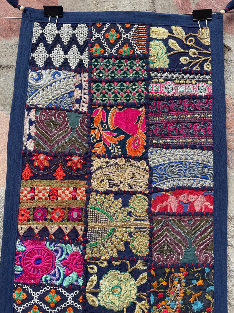 May include: A navy blue patchwork textile with intricate embroidery in various colors. The design features geometric patterns, floral motifs, and ornate details. The textile is rectangular and appears to be a wall hanging or decorative piece.