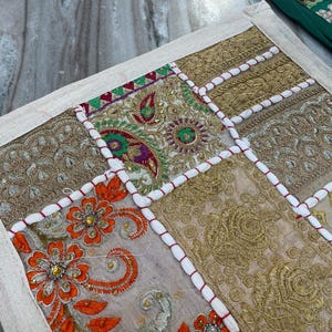 May include: A patchwork textile with a cream-colored border. The design features squares of embroidered fabric in gold, orange, and beige, with white and red cord accents. The textile is likely a decorative item, such as a pillow or wall hanging.