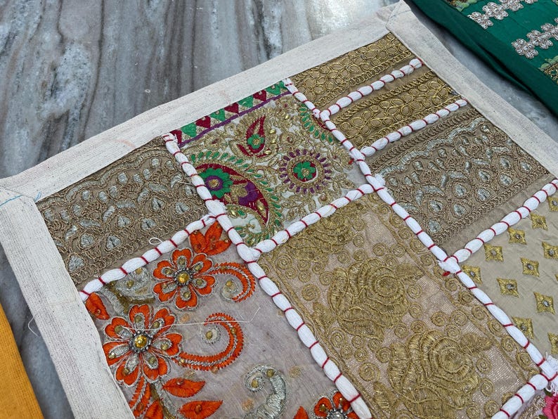 May include: A patchwork textile with a cream-colored border. The design features squares of embroidered fabric in gold, orange, and beige, with white and red cord accents. The textile is likely a decorative item, such as a pillow or wall hanging.