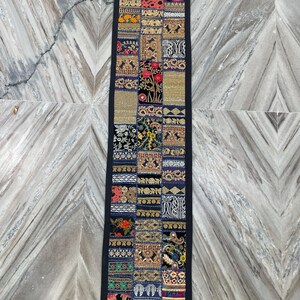 May include: A rectangular, patchwork textile with a black border. The design features a variety of embroidered squares in shades of gold, blue, red, and black. The textile is likely a decorative table runner or wall hanging.