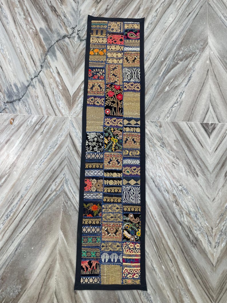 May include: A rectangular, patchwork textile with a black border. The design features a variety of embroidered squares in shades of gold, blue, red, and black. The textile is likely a decorative table runner or wall hanging.