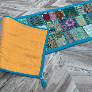 May include: A colorful patchwork table runner with a turquoise border and tassels. The runner features a variety of patterned fabric squares in shades of blue, green, red, and gold. The folded section is a solid gold color with a turquoise border.