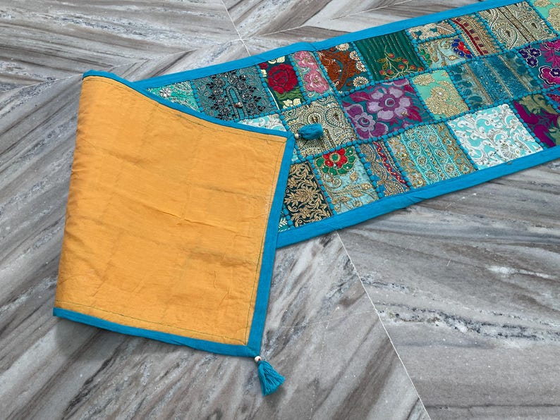 May include: A colorful patchwork table runner with a turquoise border and tassels. The runner features a variety of patterned fabric squares in shades of blue, green, red, and gold. The folded section is a solid gold color with a turquoise border.