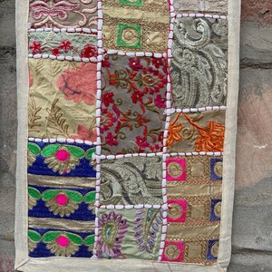 May include: A patchwork textile with a cream-colored border. The squares feature embroidered floral and geometric patterns in shades of gold, pink, blue, and green. White stitching outlines each square.