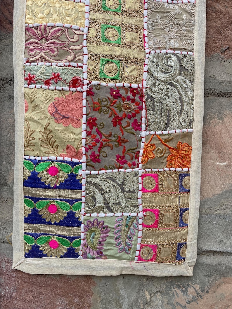 May include: A patchwork textile with a cream-colored border. The squares feature embroidered floral and geometric patterns in shades of gold, pink, blue, and green. White stitching outlines each square.