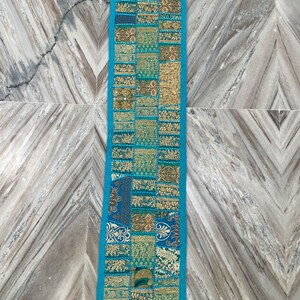 May include: A rectangular, patchwork table runner with a turquoise border. The center features a mosaic of gold, teal, and brown patterned fabric squares. The runner is likely made of a decorative textile.