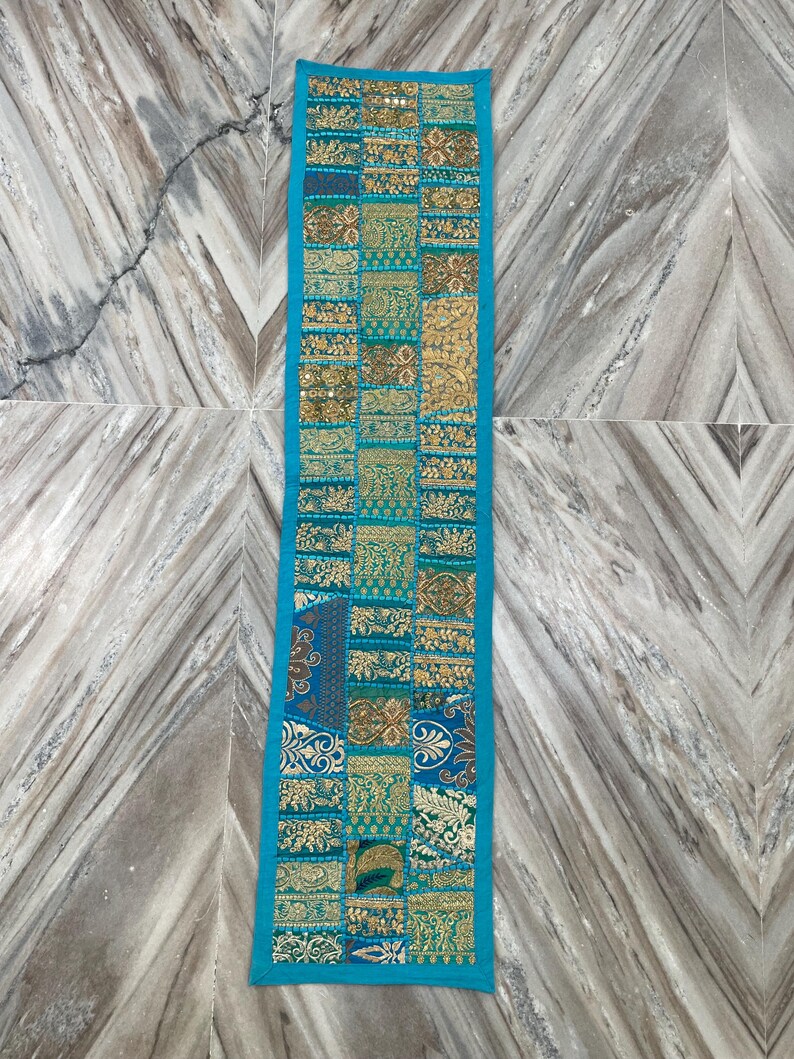 May include: A rectangular, patchwork table runner with a turquoise border. The center features a mosaic of gold, teal, and brown patterned fabric squares. The runner is likely made of a decorative textile.