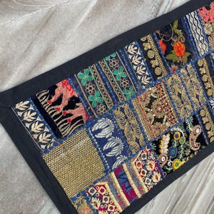 May include: A rectangular, patchwork textile with a black border. The patchwork features various embroidered patterns in gold, red, blue, and green. The textile is likely a decorative item, such as a table runner or wall hanging.