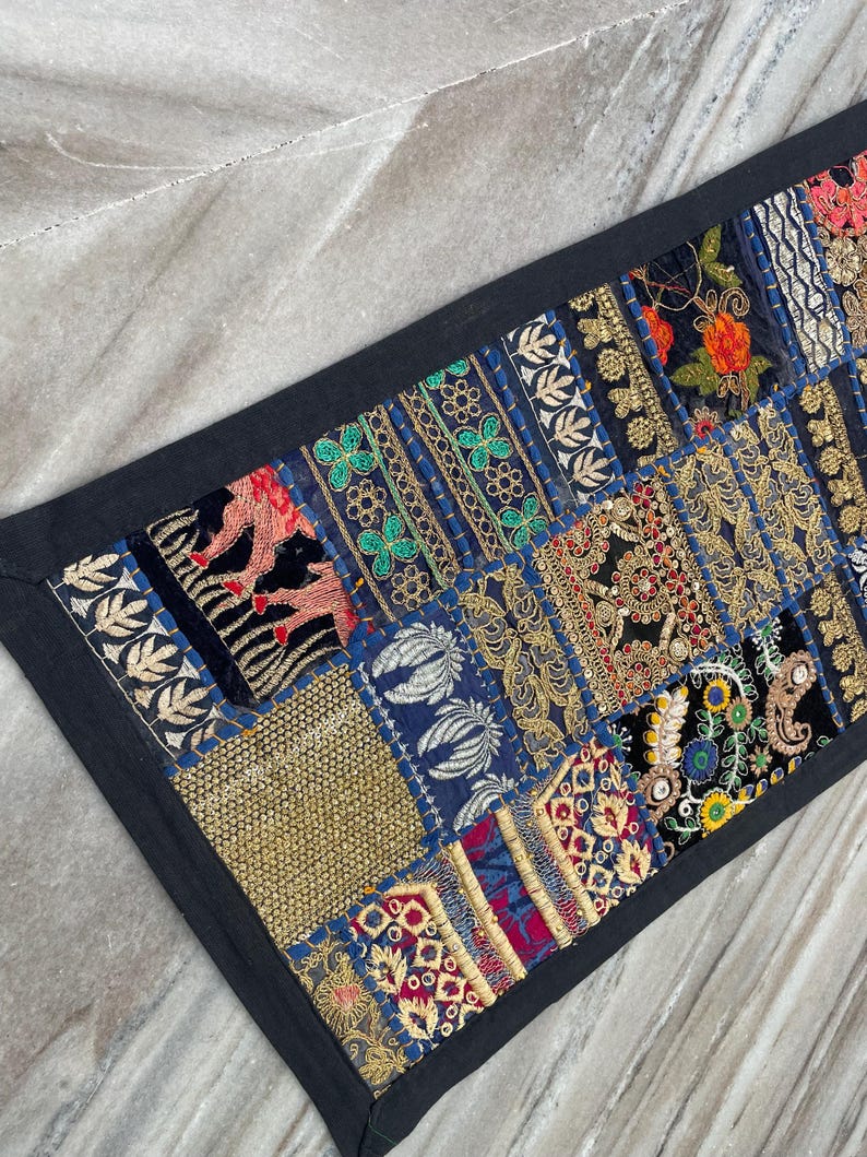 May include: A rectangular, patchwork textile with a black border. The patchwork features various embroidered patterns in gold, red, blue, and green. The textile is likely a decorative item, such as a table runner or wall hanging.
