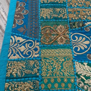 May include: A patchwork textile with a teal border and a variety of embroidered squares. The squares feature ornate gold and silver floral designs on a teal background. The textile is likely a decorative item, such as a table runner or wall hanging.