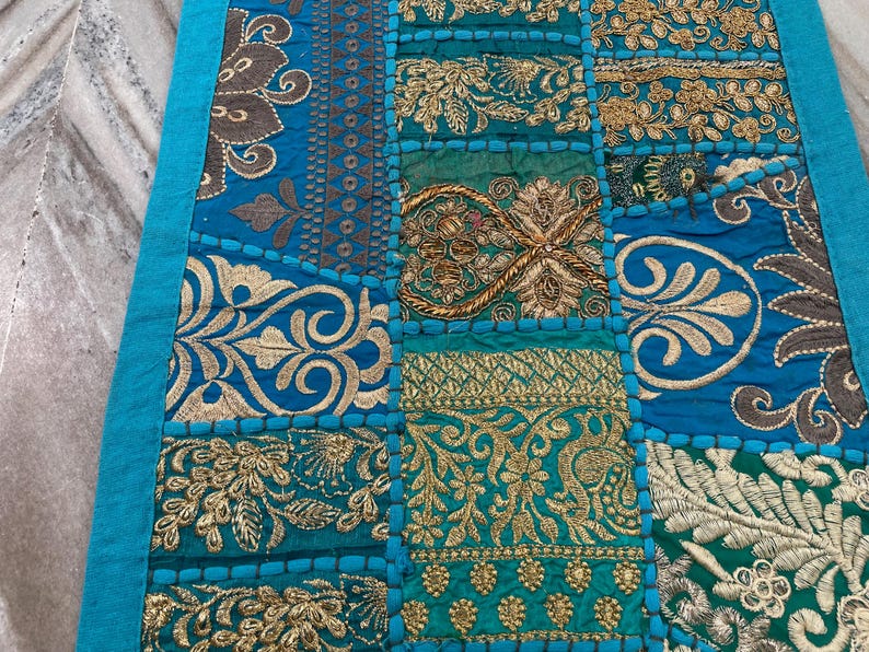 May include: A patchwork textile with a teal border and a variety of embroidered squares. The squares feature ornate gold and silver floral designs on a teal background. The textile is likely a decorative item, such as a table runner or wall hanging.