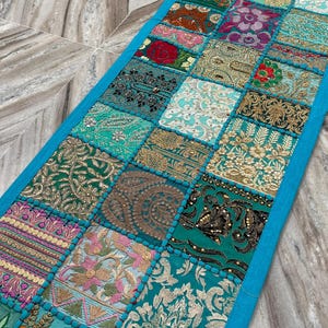 May include: A rectangular, patchwork table runner with a vibrant mix of patterned squares in teal, gold, pink, and red. The squares are bordered by a bright blue trim. The runner is made of fabric and has a decorative, bohemian style.