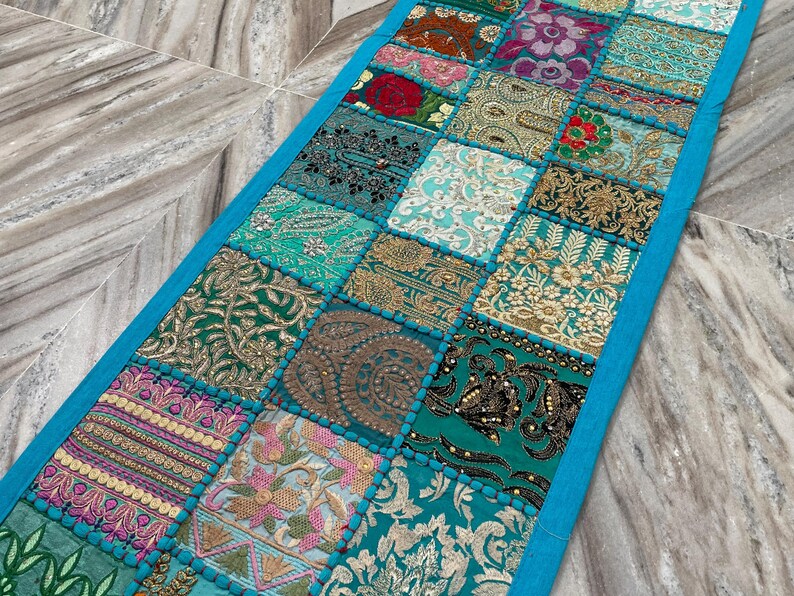 May include: A rectangular, patchwork table runner with a vibrant mix of patterned squares in teal, gold, pink, and red. The squares are bordered by a bright blue trim. The runner is made of fabric and has a decorative, bohemian style.