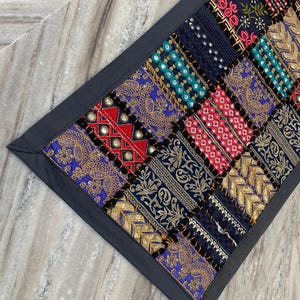 May include: A patchwork textile with a dark gray border. The design features a variety of colorful, patterned squares in shades of blue, red, gold, and teal. The textile appears to be made of multiple fabrics with embroidered details.