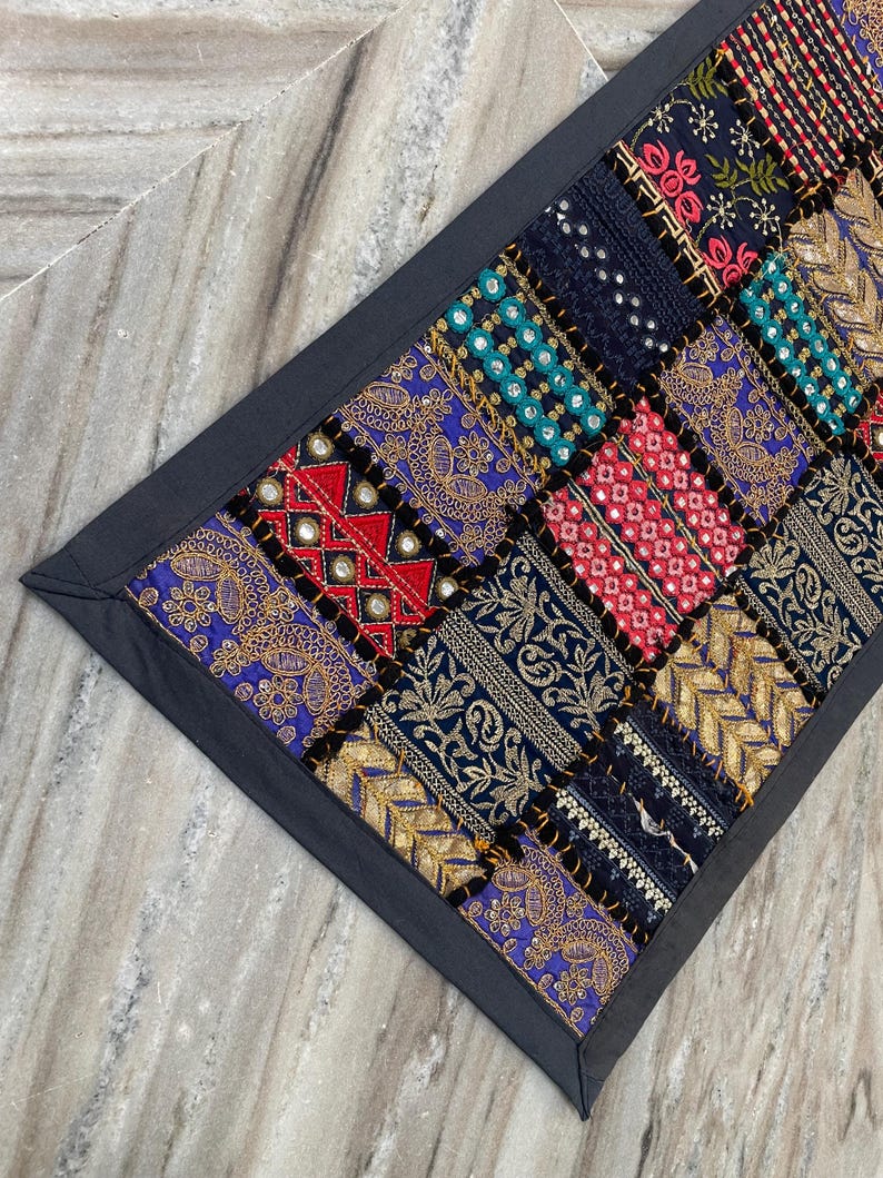 May include: A patchwork textile with a dark gray border. The design features a variety of colorful, patterned squares in shades of blue, red, gold, and teal. The textile appears to be made of multiple fabrics with embroidered details.
