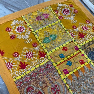 May include: A patchwork textile with a yellow border and a variety of patterned squares. The squares feature embroidered floral designs in red, white, and gold, with accents of blue and green. The textile is likely a decorative item.