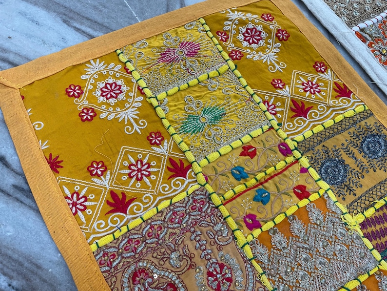 May include: A patchwork textile with a yellow border and a variety of patterned squares. The squares feature embroidered floral designs in red, white, and gold, with accents of blue and green. The textile is likely a decorative item.
