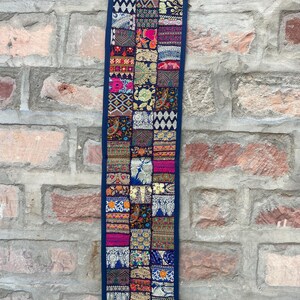 May include: A rectangular, patchwork wall hanging with a dark blue border. The patchwork is composed of various fabric squares in vibrant colors like pink, blue, and gold, with intricate patterns. The hanging is adorned with tassels at the bottom and top.
