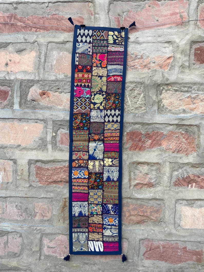 May include: A rectangular, patchwork wall hanging with a dark blue border. The patchwork is composed of various fabric squares in vibrant colors like pink, blue, and gold, with intricate patterns. The hanging is adorned with tassels at the bottom and top.